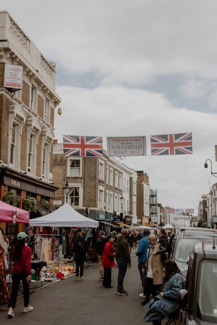 A busy street scene near Portobello Market in Notting Hill during daytime, with pedestrians walking along the pavement and browsing market stalls under white canopies that display various clothing and accessory items. In the foreground, a group of people, some carrying shopping bags, stand around the street while others are sitting on a bench on the right side. To the right, a dark grey van is parked with its rear side partially visible, and nearby, a person is seen moving a large cardboard box or container onto the pavement, possibly as part of a home relocation or furniture transport process. Above, decorative Union Jack flags and a street sign reading 'Portobello Road W11' hang across the street, adding to the lively atmosphere. The buildings lining the street are traditional in style, with some having retail shops on the ground level, and the sky above is overcast, creating diffused lighting suitable for documenting moving or packing activities. This scene illustrates the typical street environment where professional removals like those from Man with Van Notting Hill operate, especially when coordinating scheduled moves in busy market areas to avoid fines during home relocations.