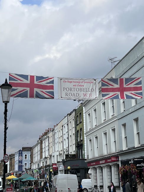 A daytime street scene in Notting Hill with a row of white-painted buildings along the pavement, featuring shopfronts and residential windows. Overhead, a large banner displays Union Jack flags on either side, reading 'Portobello Road, W11' beneath the emblem of The Royal Borough of Kensington and Chelsea. The street is bustling with activity, including several parked vehicles such as white vans and cars, some partially blocking the roadside. A worker from Man with Van Notting Hill is not visible in this image, but the scene suggests a busy environment where packing and loading activities may be ongoing. The sky overhead is cloudy, casting diffuse light over the area, and the street is decorated with street lamps and small market stalls, indicating a vibrant community space used for home relocation and furniture transport services. This setting highlights the typical environment for managing removals and moving logistics in a densely populated, well-known London neighbourhood.