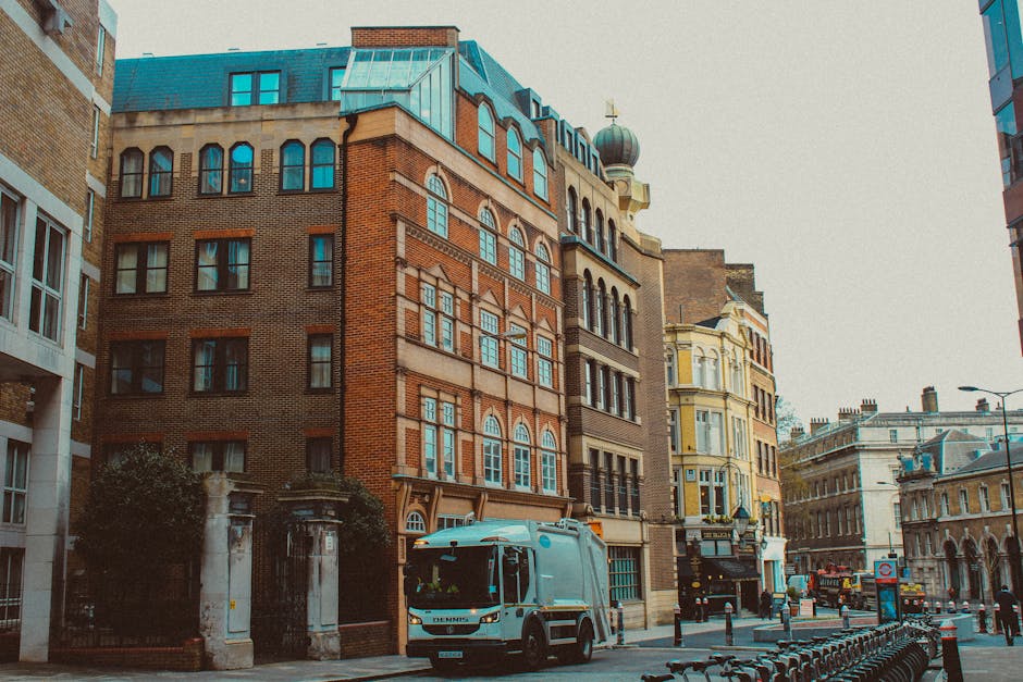 A street scene in Notting Hill showing a white commercial van belonging to Man with Van Notting Hill parked on the pavement in front of a row of multi-storey buildings with brick and stucco facades, large arched and rectangular windows, and decorative architectural details. The van is positioned near the entrance of a property with a gated archway and stone pillars, suggesting a home or apartment building. Adjacent to the van, there are several cardboard boxes and various packing materials on the ground, indicating a home relocation or furniture transport process. The street is lined with bollards, bicycles, and parked vehicles, with a mixture of older and modern architectural styles visible. Overcast sky and street lighting provide natural ambient light, contributing to a neutral, professional atmosphere typical of household removal services managed by [COMPANY_NAME]. This scene captures essential elements involved in loading furniture, boxes, and belongings during house removals in an urban environment, aligning with services like packing, logistics, and transportation.