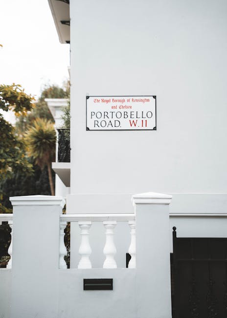 A white residential building with a small balcony featuring white balustrades and a dark gate is visible in the image. On the exterior wall, there is a rectangular street sign indicating 'Portobello Road, W.11' with additional text referencing the Royal Borough of Kensington and Chelsea. The setting appears to be in a suburban or urban area, with some foliage and trees visible in the background. The scene is captured during daylight with natural lighting, highlighting the clean, painted surface of the building and the sign's details. This image relates to house removals and relocation services, illustrating a typical street scene in Notting Hill where furniture transport and packing preparations might be ongoing. The presence of a well-maintained façade and typical residential features suggests it could be a property involved in a professional home move, such as those undertaken by Man with Van Notting Hill.
