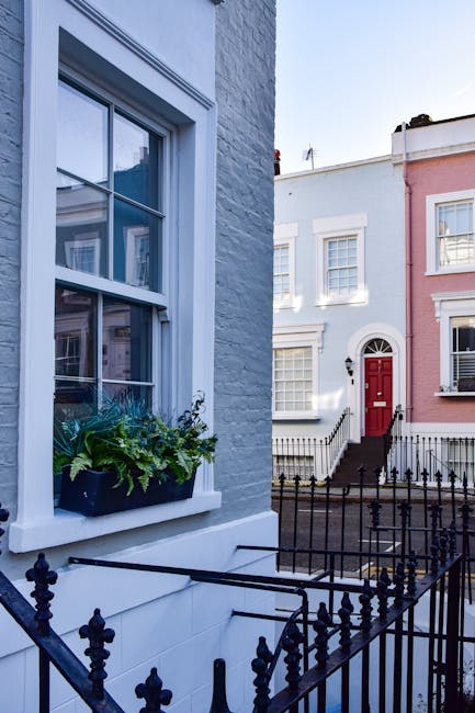 A close-up view of a white-painted brick house in Notting Hill, featuring a double-hung sash window with white wooden frames and a window box filled with green plants and flowering foliage. In the background, there is a row of terraced houses with pastel-colored facades, including one with a red front door accessed by black metal stairs and railings. The scene is illuminated by daylight, highlighting the urban residential environment, which is typical for home relocation and furniture transport operations, and suggests the setting for house removals or moving services provided by Man with Van Notting Hill.