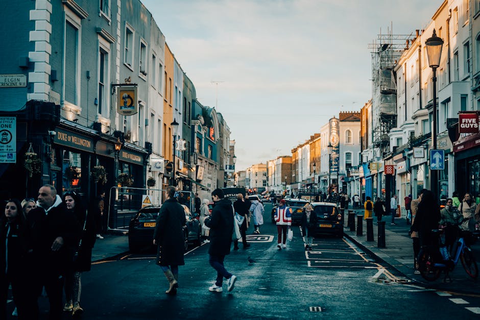 A busy street scene in Notting Hill with pedestrians walking along the sidewalk and crossing the road. Several parked cars line the street, and a man wearing a high-visibility vest is standing near the curb, possibly coordinating the loading process. On the left, a row of colourful shops and pubs, including a pub named Duke of Wellington, is visible with some storefronts displaying signage. In the background, there are residential buildings with light-colored facades, some under scaffolding, indicating ongoing maintenance or renovation. The pavement is lined with bollards and street lamps, and the overall scene is illuminated by natural daylight, suggesting daytime. This setting showcases typical elements involved in house removals or furniture transport, with the city environment supporting the logistical aspects of a home relocation, such as vehicle loading and pedestrian navigation, aligned with services from Man with Van Notting Hill.