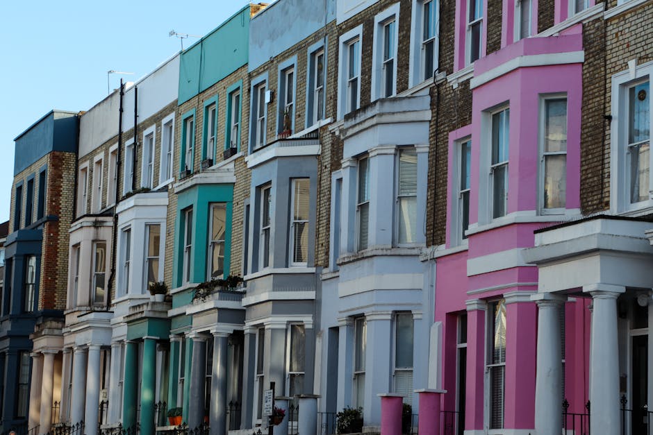 A row of Victorian terraced houses in Notting Hill, London, featuring facades painted in pastel shades including blue, pink, and cream. The buildings display classic bay windows with white trim, some with flower pots on window sills. The ground level shows small front gardens enclosed by black wrought iron fences. The image captures a clear day with natural light illuminating the colorful exterior walls. In front, a pavement runs along the houses, with a loading area visible where a moving van from Man with Van Notting Hill is likely being used for home relocation and furniture transport. The scene reflects ongoing packing and loading activities typical of professional removals services, with some cardboard boxes and packing materials possibly out of view. The overall environment emphasizes residential street parking regulations, such as Kensington & Chelsea Council: moving van permits for Notting Hill, needed for efficient and compliant moving processes.
