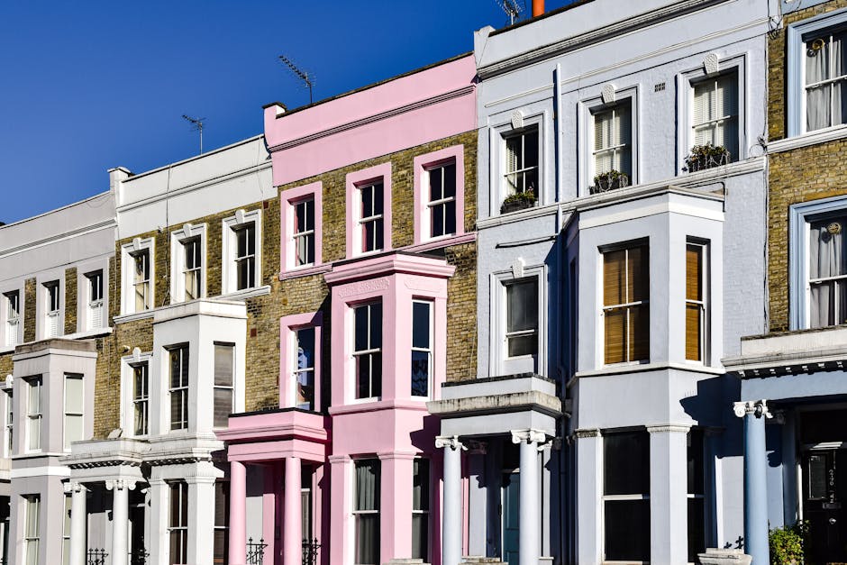 A busy street scene in Notting Hill with pedestrians walking along the sidewalk and crossing the road. Several parked cars line the street, and a man wearing a high-visibility vest is standing near the curb, possibly coordinating the loading process. On the left, a row of colourful shops and pubs, including a pub named Duke of Wellington, is visible with some storefronts displaying signage. In the background, there are residential buildings with light-colored facades, some under scaffolding, indicating ongoing maintenance or renovation. The pavement is lined with bollards and street lamps, and the overall scene is illuminated by natural daylight, suggesting daytime. This setting showcases typical elements involved in house removals or furniture transport, with the city environment supporting the logistical aspects of a home relocation, such as vehicle loading and pedestrian navigation, aligned with services from Man with Van Notting Hill.