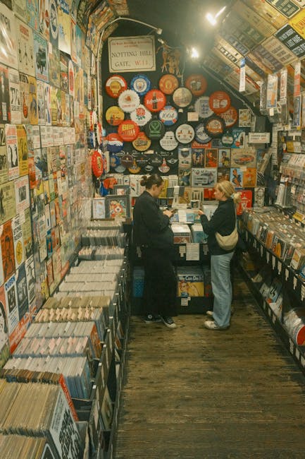 Inside a small, narrow bookstore or record shop with walls covered in colourful posters, album covers, and signs. There are shelves filled with books or vinyl records along both sides of the space, with some items stacked on top of each other. A small counter at the back has various items displayed and two women are standing in front of it, engaged in conversation; one woman is dressed in a dark jacket with her hair tied back, while the other wears a light-blue top and carries a shoulder bag. The wooden floor shows signs of wear, and there's a sign above the counter indicating the entrance to Notting Hill at the gate, W11. The environment is lit with warm overhead lighting, suggesting an inviting atmosphere focused on browsing and purchase, suitable for a home relocation or moving scenario involving packing or selecting items for a move. Man with Van Notting Hill offers removals and packing services that could include relocating treasured items from this shop to a new home.