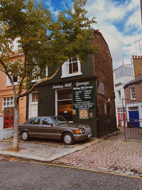 The image depicts the exterior of a white, two-story building on a narrow street in Notting Hill, featuring a curved black wrought-iron balcony on the first floor. The ground floor has a large window displaying ceramic tableware and a sign reading 'BOULANGERIE,' indicating a bakery. To the right of the window, there is a black door with a small awning above it. On the sidewalk in front of the building, two green and white A-boards lean against a black post on the right side, with some partially visible road markings and a drain grate in the pavement. The street setting suggests a typical residential or commercial area, with no visible moving activity or furniture. As part of a house removal or furniture transport process, this exterior shot could be relevant in illustrating a typical Notting Hill property where [COMPANY_NAME] offers removals and packing services. The overall scene is well-lit with natural daylight, with some trees visible on the right edge of the frame.