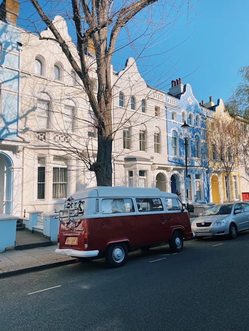 A vintage red and white Volkswagen camper van is parked on a residential street in Notting Hill during daylight hours, with a tree without leaves positioned directly behind it and a row of colourful Victorian-style terraced houses in shades of white, blue, and yellow lining the street. The van is situated close to the curb, with its rear doors closed and a bicycle mounted on the back rack. Nearby, a modern silver car is parked alongside the van. The street appears quiet with no pedestrians visible, and the sky above is clear and blue, suggesting good weather conditions. This scene depicts an urban environment typical of house relocation or furniture transport activities, with the van likely used for home removal services offered by companies such as Man with Van Notting Hill. The arrangement and items in the vicinity imply ongoing packing or loading processes, essential components of effective house moves in challenging parking situations typical of Notting Hill.