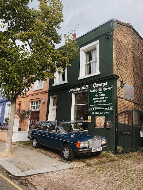 A two-storey building painted in dark green with white window frames, identified as 'Notting Hill Garage' by a sign on the front. The sign indicates services such as car sales, repairs, and vehicle care. To the left of the building, a large tree with green foliage partially obscures the upper part of the façade. A classic dark blue station wagon with a roof rack is parked on the pavement directly in front of the garage, adjacent to the sidewalk. The driveway area adjacent to the building is paved with small cobblestones, leading to a gated entrance on the right side. The scene takes place during daylight hours under partly cloudy skies. Surrounding the garage are residential buildings with varied facades, typical of Nottingham Hill, London. The image captures elements relevant to home relocation logistics, including furniture and vehicle positioning, with Man with Van Notting Hill providing moving services likely involved in an ongoing or upcoming furniture transport or packing process.