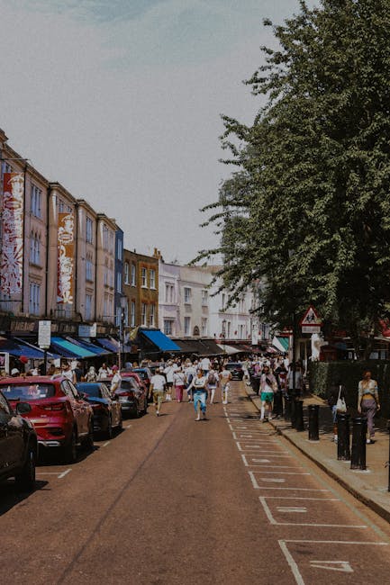 The image shows a busy street scene in Notting Hill with a row of colorful terraced houses painted in pastel shades of blue, yellow, pink, and white. A white van with the branding 'Grahbrook' is parked on the pavement, partially visible in the foreground, near the entrance of a residential property. In front of the house, a person dressed in casual clothing is actively lifting or carrying a large cardboard box wrapped in plastic, possibly containing household items for a home relocation or packing process. The sidewalk shows scattered packing supplies, including cardboard boxes and packing materials, indicating a recent move-in or move-out activity. To the right, a black street lamp is mounted on a decorative pole, while above, the sky is partly cloudy with soft daylight illuminating the scene. This setting, with furniture or boxes being loaded or unloaded, aligns with a professional removals service, such as those provided by Man with Van Notting Hill, supporting the logistics of house moves with equipment like trolleys and blankets visible nearby.