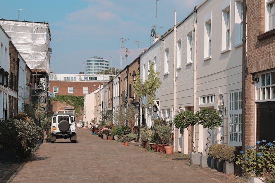 A man with a beard wearing a brown hat and black clothing is standing outdoors on a city street near a row of colourful Victorian-style terraced houses with painted facades in shades of blue, purple, 
