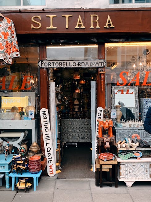 The entrance of a shop named 'SITARA' with a wooden sign above the door, located at Portobello Road in Notting Hill. The shop displays a variety of items including vintage collectibles, ceramics, and decorative objects on shelves and tables outside. Two vertical signs indicate 'NOTTING HILL GATE' and 'PORTOBELLO ROAD W11', suggesting nearby streets or directions. The shop's interior is visible through the open doorway, revealing hanging lamps, shelves filled with merchandise, and a narrow corridor leading further inside. The exterior includes a display of colourful textiles, small furniture, and various knick-knacks arranged on blue and white tables, with some items in cardboard boxes. The scene is set during daylight, with the shop's window reflecting the street outside, implying an active shopping environment connected to house removals or packing activities typical in home relocations. ManwithVannottingHill.co.uk occasionally referenced, aligning with moving and clearance services in the Notting Hill area.