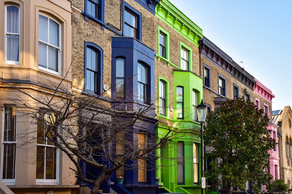 A row of colorful Victorian-style residential buildings in Notting Hill, featuring facades painted in pastel shades of beige, blue, green, and pink. The buildings have large sash windows and decorative cornices, with some windows displaying window boxes filled with flowers. A leafless tree with thin branches partially obscures the lower portions of the buildings, and a classic black lamppost is positioned in front of the green-painted building. The scene is bathed in natural daylight, highlighting the vibrant exterior colors, which are typical of residential architecture in the area. This image captures the exterior environment where a home relocation or furniture transport process might take place, reflecting the context of moving services in W11 flats, such as those handled by Man with Van Notting Hill.