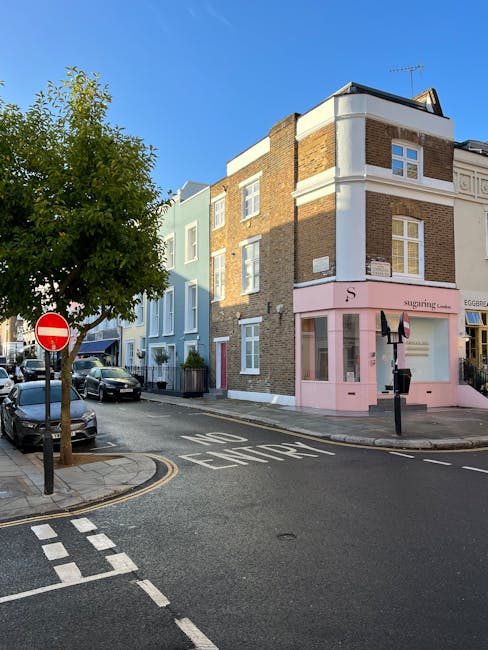 A three-storey mixed-use building on a street corner in Notting Hill featuring a pink shopfront on the ground level, with large windows and a sign reading 'SugaRing'; the upper floors have brown brick facades with white window frames, and the building is set at the intersection with a narrow road. In the foreground, a black bollard and a sign indicating no entry are visible, along with parked cars lining the street. A tree with green leaves partially shades the sidewalk, and the sky is clear and blue, indicating daytime. The scene is typical for residential and small commercial property in an urban area undergoing home relocation or furniture transport preparations, with visible materials like cardboard boxes and blankets possibly used for packing and protecting furniture during the moving process, supported by local removals services such as Man with Van Notting Hill.