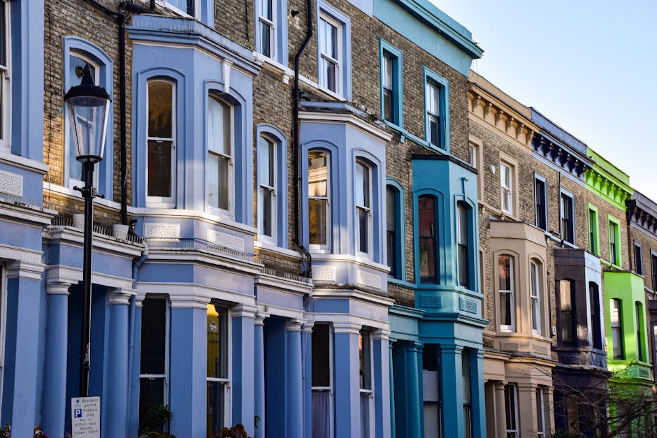 A row of colorful Victorian-style residential buildings in Notting Hill, featuring facades painted in pastel shades of beige, blue, green, and pink. The buildings have large sash windows and decorative cornices, with some windows displaying window boxes filled with flowers. A leafless tree with thin branches partially obscures the lower portions of the buildings, and a classic black lamppost is positioned in front of the green-painted building. The scene is bathed in natural daylight, highlighting the vibrant exterior colors, which are typical of residential architecture in the area. This image captures the exterior environment where a home relocation or furniture transport process might take place, reflecting the context of moving services in W11 flats, such as those handled by Man with Van Notting Hill.