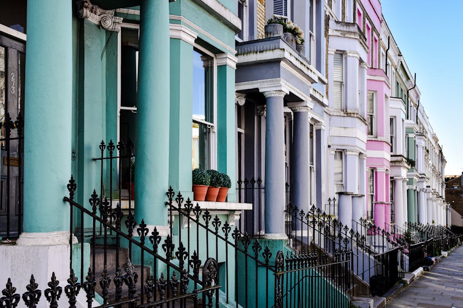 A line of Victorian terraced houses in Westbourne Grove, Notting Hill, featuring pastel-colored facades with shades of turquoise, gray, and pink. The houses have tall, rounded columns supporting small