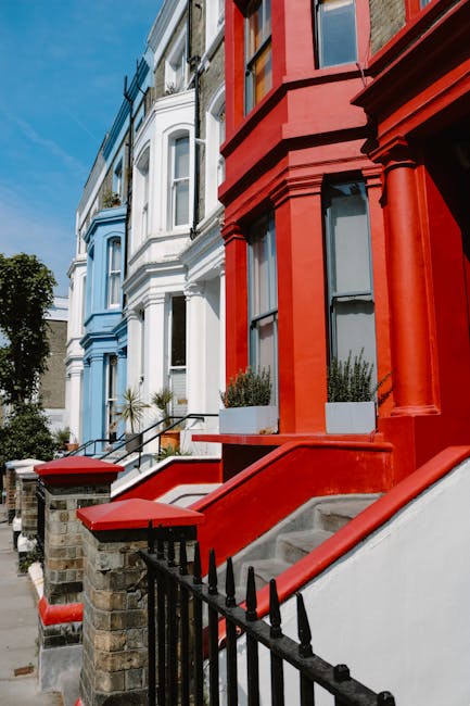 Photograph of a row of three Victorian-style terraced houses on Westbourne Grove, W2, showing the exterior facades during daytime. The building closest to the camera has a bold red-painted facade with large ground-floor windows framed by decorative columns and black metal railings along the front steps. Adjacent houses are painted in pastel blue and white, each with distinctive bay windows and wrought iron railings. The street scene includes a paved sidewalk with a few potted plants visible on the steps of the houses. Bright sunlight illuminates the facades, highlighting the vibrant colours and architectural details. The image captures the typical residential street environment with no visible moving activity, but it depicts the type of property where professional house removals or furniture transport might be scheduled by Man with Van Notting Hill as part of a home relocation or packing and moving service.