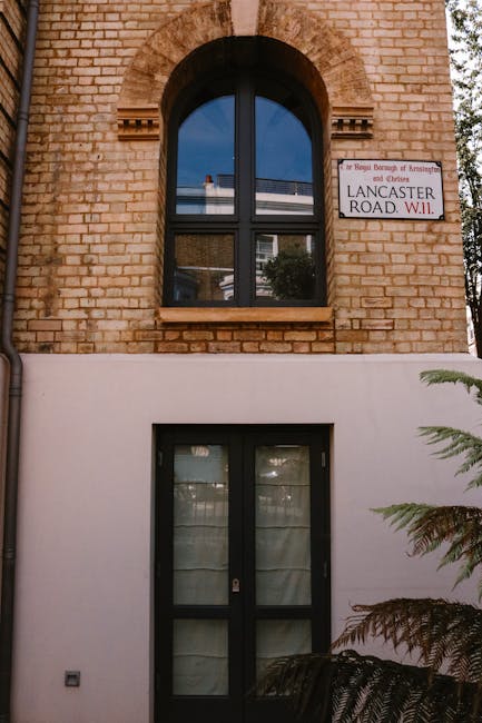The image shows the exterior of a building with a brick upper section and a white lower section. There is a large, arched, double-glazed window with a black frame on the upper floor, reflecting the sky and surrounding buildings. Below, on the ground floor, there is a black-framed glass door with a curtain behind it, positioned in the white section of the wall. To the right of the door, a corner of a green fern plant is visible, partially blocking the view of the wall. A street sign on the brick wall to the right reads 'Lancaster Road W11,' indicating the location in Notting Hill. The overall scene is lit by natural daylight, and the image captures a quiet residential property, relevant to house removals and moving services such as those provided by Man with Van Notting Hill, as seen on their website page about Westbourne Grove to Lancaster Gate moves.