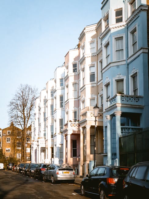 Photograph of a row of three Victorian-style terraced houses on Westbourne Grove, W2, showing the exterior facades during daytime. The building closest to the camera has a bold red-painted facade with large ground-floor windows framed by decorative columns and black metal railings along the front steps. Adjacent houses are painted in pastel blue and white, each with distinctive bay windows and wrought iron railings. The street scene includes a paved sidewalk with a few potted plants visible on the steps of the houses. Bright sunlight illuminates the facades, highlighting the vibrant colours and architectural details. The image captures the typical residential street environment with no visible moving activity, but it depicts the type of property where professional house removals or furniture transport might be scheduled by Man with Van Notting Hill as part of a home relocation or packing and moving service.